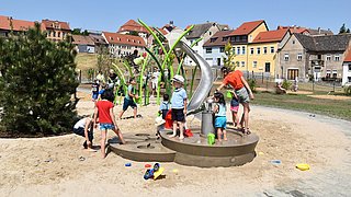 Foto, Kinder spielen an einem Wasserspielplatz mit Rutsche und Sand in einem Wohngebiet mit bunten Häusern im Hintergrund