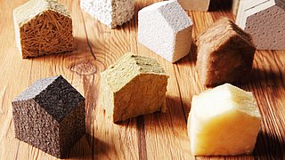 Foto, mehrere Materialproben verschiedener Baustoffe in Form eines kleinen Hauses, nebeneinander aufgestellt auf einem Tisch.