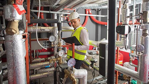 Foto, Person mit Sicherheitshelm, Klemmbrett und Warnweste steht in einem Raum mit Heizungstechnik und macht Notizen.