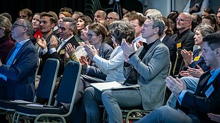 Foto, mehrere Personen sitzen in Reihen auf Stühlen und klatschen, einige tragen Anstecknadeln, die Kleidung ist überwiegend formell.