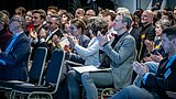 Foto, mehrere Personen sitzen in Reihen auf Stühlen und klatschen, einige tragen Anstecknadeln, die Kleidung ist überwiegend formell.