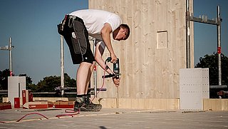 Foto, eine Person in Arbeitskleidung bohrt auf einer Baustelle in den Fußboden.