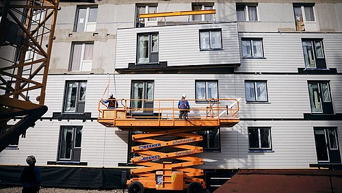 Foto, zwei Bauarbeiter auf einer orangefarbenen Hebebühne vor einem mehrstöckigen Gebäude mit weißen Fassadenelementen