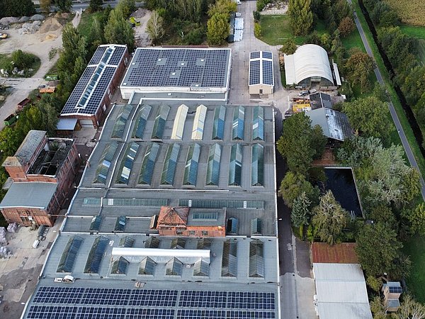 Luftaufnahme des Geländes Alte Filzfabrik mit mehreren großen Fabrikgebäuden, Solarpaneelen auf den Dächern und umliegenden Bäumen und Feldern.