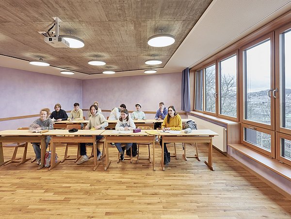 Foto, Schulklasse mit zehn Schülern sitzt an Tischen in einem hellen Klassenraum mit großen Fenstern und Holzboden