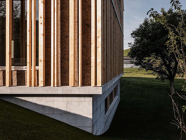 Foto, Ecke eines Gebäudes mit Korkdämmung zwischen vertikalen Holzlatten auf einem Betonsockel neben einem Baum und Rasenfläche