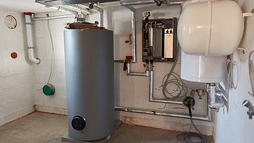 Foto, ein Heizungsraum mit einem großen, silbernen Warmwasserspeicher in der Mitte. An der Wand sind Rohre und ein Heizungsregler sichtbar. Rechts steht ein weißer Ausdehnungsbehälter.