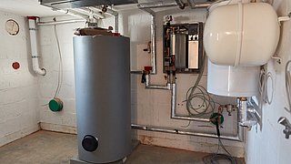 Foto, ein Heizungsraum mit einem großen, silbernen Warmwasserspeicher in der Mitte. An der Wand sind Rohre und ein Heizungsregler sichtbar. Rechts steht ein weißer Ausdehnungsbehälter.