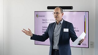 Foto, Mann mit Brille und Sakko hält Papiere in beiden Händen und gestikuliert vor einem Bildschirm mit Text.