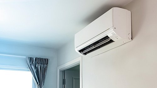 Foto, Ein weißes Klimagerät, das an einer hellen Wand montiert ist, wobei im Hintergrund ein Fenster und ein Vorhang zu sehen sind.
