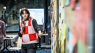 Foto, Person mit rot-weiß kariertem Strickweste und schwarzem Hemd hält Mikrofon und zeigt auf eine Pinnwand mit bunten Notizzetteln.