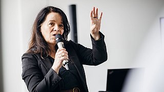 Foto, Frau in schwarzem Blazer hält Mikrofon und hebt die rechte Hand während einer Präsentation