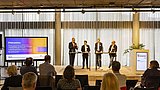Foto, eine Podiumsdiskussion mit vier Rednern an Stehtischen in einem hellen Raum mit großen Fenstern und einem gesprächsfreudigen Publikum.