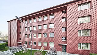 Foto, modernes rotes Gebäude mit rautenförmiger Fassade, großen Fenstern und einem Fahrradunterstand mit Fahrrädern in einer Grünanlage.