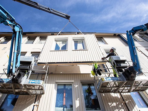 Foto, zwei Hebebühnen mit jeweils einer Person stehen vor einer Hausfassade. In der Mitte befindet sich ein Fassadenelement, was vor der Fassade hängt.