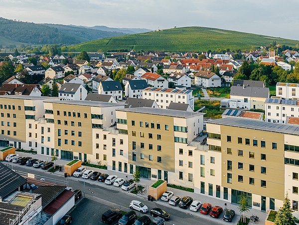 Mehrere moderne, beige-weiße Wohngebäude mit Flachdächern in Schallstadt, im Hintergrund bewaldete Hügel und weitere Wohnhäuser.