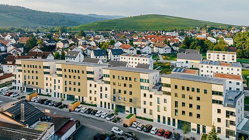 Mehrere moderne, beige-weiße Wohngebäude mit Flachdächern in Schallstadt, im Hintergrund bewaldete Hügel und weitere Wohnhäuser.
