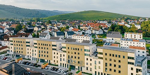 Mehrere moderne, beige-weiße Wohngebäude mit Flachdächern in Schallstadt, im Hintergrund bewaldete Hügel und weitere Wohnhäuser.