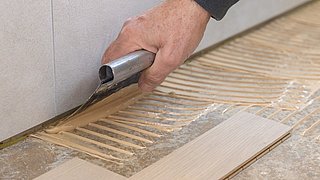 Foto, Hand mit Zahnspachtel trägt Kleber auf Boden vor Parkettverlegung auf.