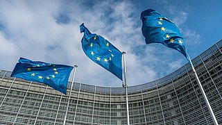 Foto, Drei EU-Flaggen mit gelben Sternen wehen vor einem modernen, gläsernen Gebäude unter einem bewölkten Himmel.