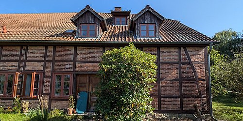 Foto, Fachwerkhaus mit braunem Ziegeldach, mehreren Fenstern und einer offenen Holztür, davor ein großer grüner Strauch