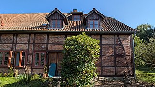 Foto, Fachwerkhaus mit braunem Ziegeldach, mehreren Fenstern und einer offenen Holztür, davor ein großer grüner Strauch
