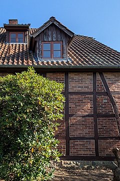 Foto, Fachwerkhaus mit braunem Ziegeldach, mehreren Fenstern und einer offenen Holztür, davor ein großer grüner Strauch