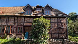 Foto, Fachwerkhaus mit braunem Ziegeldach, mehreren Fenstern und einer offenen Holztür, davor ein großer grüner Strauch