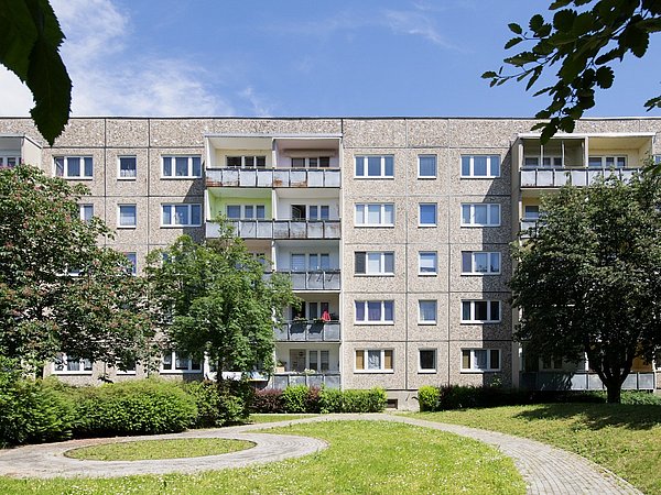 Mehrstöckiges Wohnhaus vor der Sanierung mit Balkonen, davor ein gepflegter Garten mit Rasen, Büschen und einem runden gepflasterten Weg.