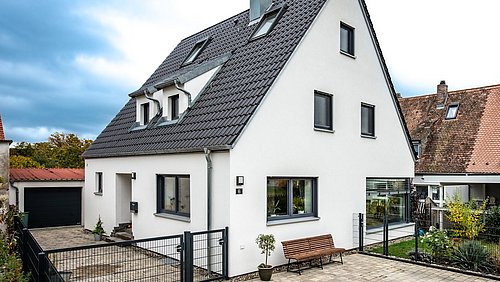 Foto, modernes weißes Haus mit Satteldach, umgeben von einem gepflasterten Innenhof und üppigem Grün, unter einem bewölkten Himmel.