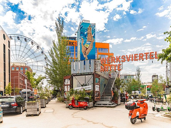 Foto, Eingang zum Werksviertel Mitte in München mit großem Schriftzug, buntem Wandbild, roten Rollern und Riesenrad im Hintergrund