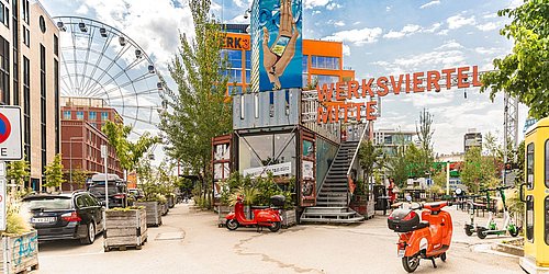 Foto, Eingang zum Werksviertel Mitte in München mit großem Schriftzug, buntem Wandbild, roten Rollern und Riesenrad im Hintergrund