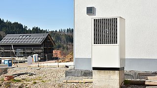 Foto, im Vordergrund ein Haus mit davorstehender Wärmepumpe, im Hintergrund ein Haus im Bau mit Solarkollektoren auf dem Dach.