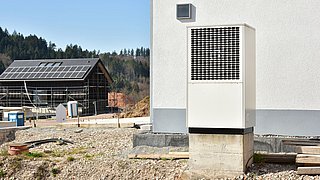 Foto, im Vordergrund ein Haus mit davorstehender Wärmepumpe, im Hintergrund ein Haus im Bau mit Solarkollektoren auf dem Dach.