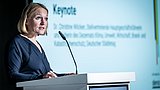 Foto, Frau mit blauer Bluse steht an einem weißen Rednerpult mit Mikrofon, im Hintergrund unscharfe Projektion.