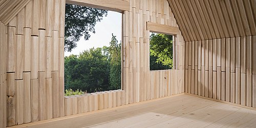 Foto, Innenraum eines Holzhauses mit zwei quadratischen Fenstern, durch die grüne Bäume sichtbar sind.