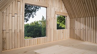 Foto, Innenraum eines Holzhauses mit zwei quadratischen Fenstern, durch die grüne Bäume sichtbar sind.