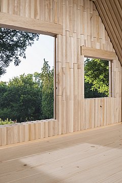 Foto, Innenraum eines Holzhauses mit zwei quadratischen Fenstern, durch die grüne Bäume sichtbar sind.