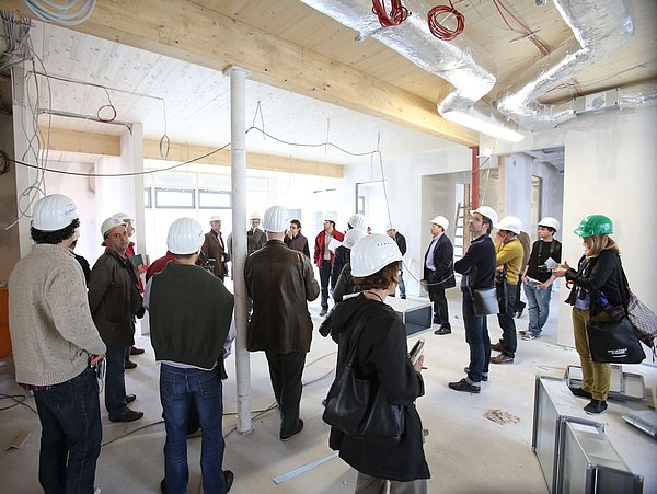 Foto, eine Gruppe Menschen auf einer Baustelle. Alle Personen tragen weiße Baustellen-Schutzhelme.