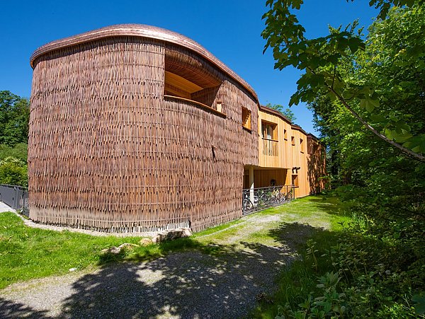 Foto, das organisch geformte Rosana Waldhaus von außen mit einer mit Holz und Weidenzweigen verkleideten Fassade.