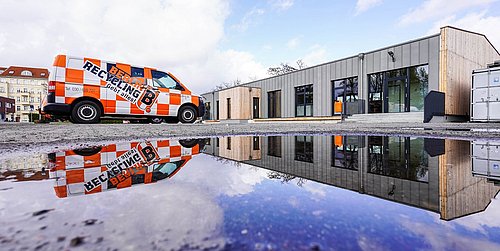 Foto, Oranger Recycling-Transporter mit Schriftzug 'Berlin Recycling' vor modernem Gebäude, Spiegelung im Wasserpfütze