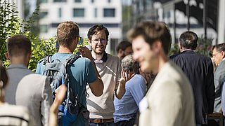 Foto, eine Gruppe von Menschen unterhält sich im Freien, umgeben von Grün, in einer modernen städtischen Umgebung, einige davon in Geschäftskleidung.
