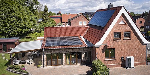 Foto, Zweistöckiges rotes Backsteinhaus mit Solarpaneelen auf dem Dach, einer Wärmepumpe vor dem Gebäude und einem überdachten Sitzbereich im Garten.
