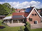 Foto, Zweistöckiges rotes Backsteinhaus mit Solarpaneelen auf dem Dach, einer Wärmepumpe vor dem Gebäude und einem überdachten Sitzbereich im Garten.
