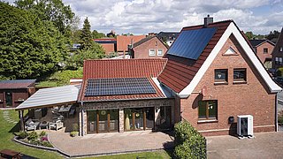 Foto, Zweistöckiges rotes Backsteinhaus mit Solarpaneelen auf dem Dach, einer Wärmepumpe vor dem Gebäude und einem überdachten Sitzbereich im Garten.