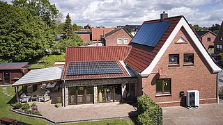 Foto, Zweistöckiges rotes Backsteinhaus mit Solarpaneelen auf dem Dach, einer Wärmepumpe vor dem Gebäude und einem überdachten Sitzbereich im Garten.
