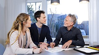 Foto, drei Personen sitzen an einem Tisch mit Dokumenten und diskutieren in einem hellen Raum mit großen Fenstern und zwei Hängeleuchten.