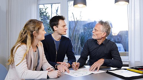 Foto, drei Personen sitzen an einem Tisch mit Dokumenten und diskutieren in einem hellen Raum mit großen Fenstern und zwei Hängeleuchten.