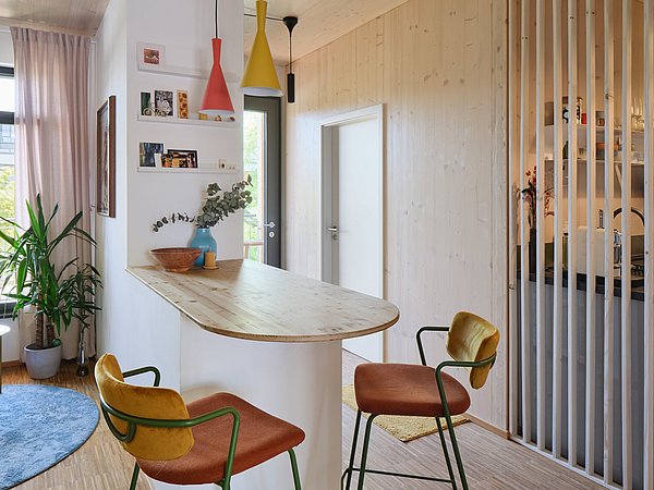 Foto, Moderner Wohnraum mit Holztisch, zwei Stühlen mit orangefarbenen Sitzflächen, Pflanzen und offener Küche mit weißen Lamellen als Raumteiler