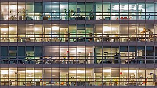 Foto, frontaler Blick auf die vollständig verglaste Fassade eines Bürogebäudes bei Dunkelheit. In den beleuchteten Räumen sind Einrichtungsgegenstände zu erkennen.